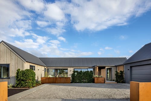 Modern single-storey home arranged around a sunlit central courtyard, with living spaces, landscaped garden beds and a garage opening onto a gravel forecourt beneath a bright blue sky.