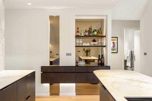 Modern Clevedon show-home kitchen with dark timber cabinetry, pale stone benchtops, a large island, integrated appliances and open shelving.